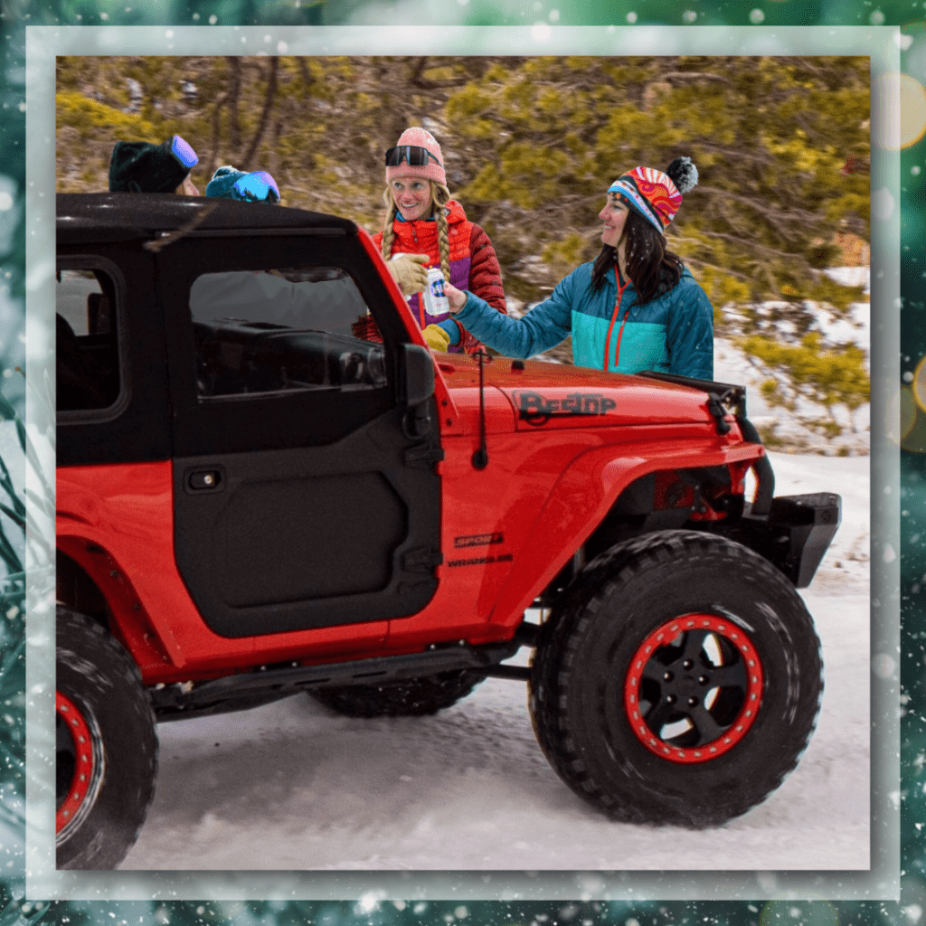 Group of friends gathered beside a red Jeep Wrangler in the snow, wearing colorful winter jackets and hats while sharing drinks and chatting