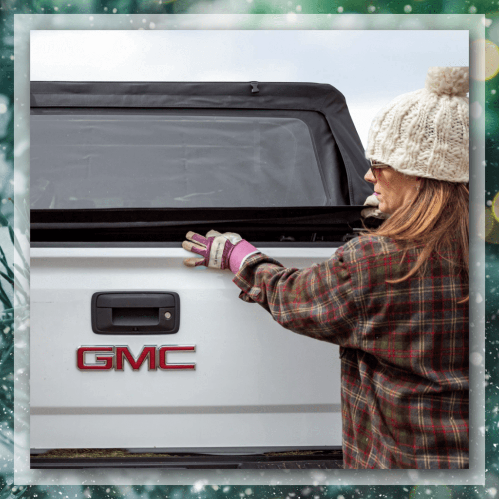 Woman in a plaid jacket and knit beanie closing the tailgate of a white GMC truck equipped with a Softopper, set against a cold outdoor backdrop